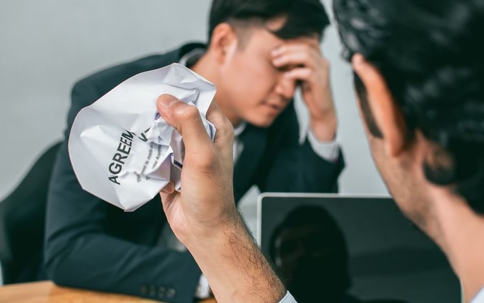 Two businessmen, one holding a crumpled agreement, looking stressed during a discussion